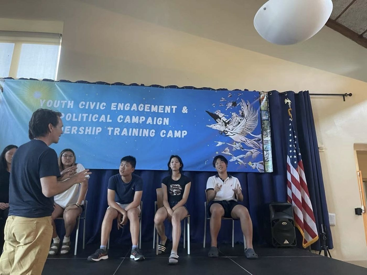 A trainer standing and speaking to students seated on a low stage, under a blue banner reading Youth Civic Engagement and Political Campaign Leadership Training Camp.