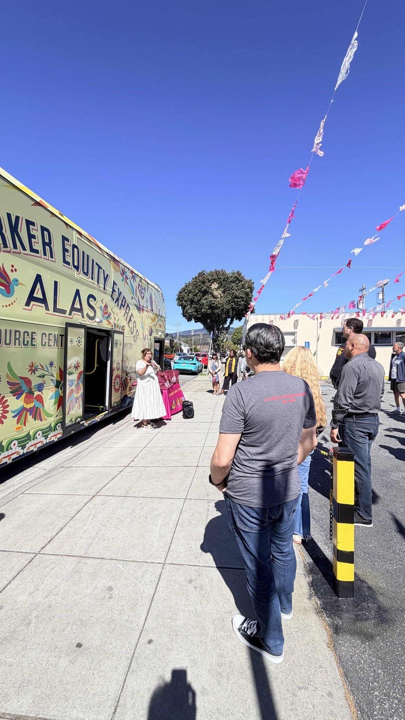 A woman in a white dress speaking to a small group on a sunny pavement beside a long bus painted with flowers and birds and lettered Farmworker Equity Express, ALAS, Mobile Resource Center, with paper banners strung overhead.